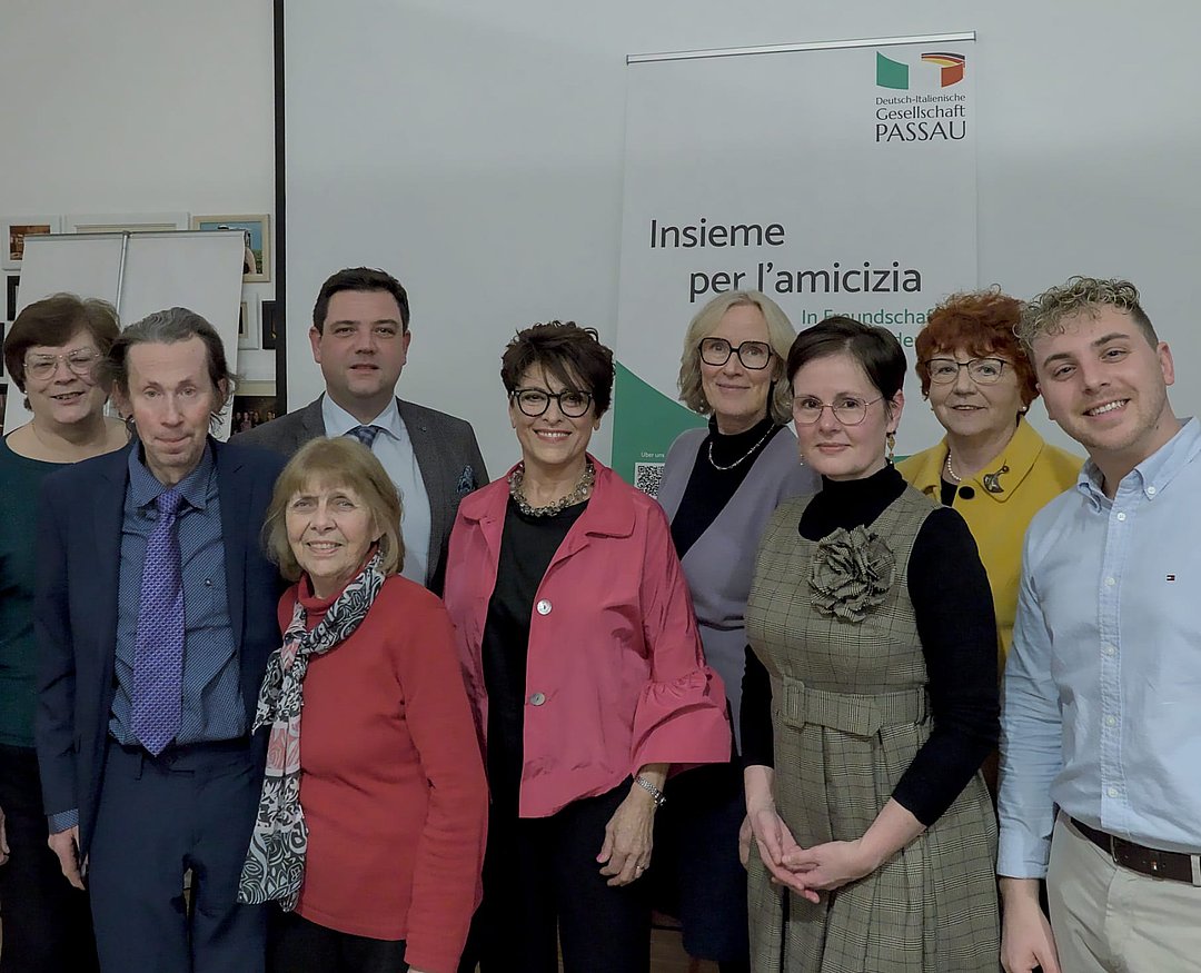 Eine Gruppe von neun Erwachsenen, Männern und Frauen, steht in einem Innenraum zusammen und lächelt in die Kamera. Auf einem Banner hinter ihnen steht Insieme per lamicizia mit dem Logo der Deutsch-Italienischen Gesellschaft Passau.