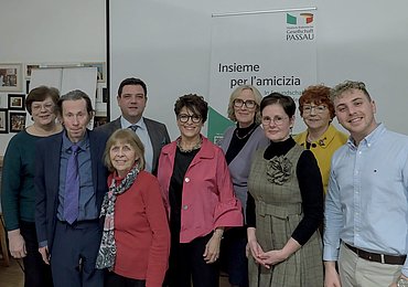 Eine Gruppe von neun Erwachsenen, Männern und Frauen, steht in einem Innenraum zusammen und lächelt in die Kamera. Auf einem Banner hinter ihnen steht Insieme per lamicizia mit dem Logo der Deutsch-Italienischen Gesellschaft Passau.