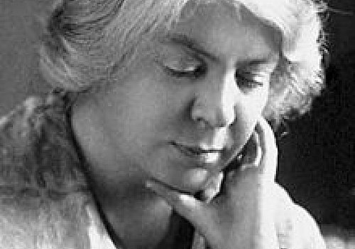 Eine Frau mit hellem Haar blickt nachdenklich nach unten und stützt ihr Kinn auf ihre Hand. Sie trägt ein gemustertes Tuch über den Schultern. Das Foto ist in schwarz-weiß gehalten.